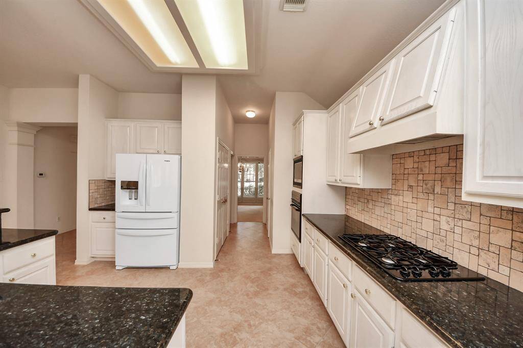 This kitchen features sleek black countertops, white cabinetry, a built-in gas stove, and a matching double oven. It includes a spacious refrigerator and a stylish tiled backsplash, offering both functionality and modern appeal.