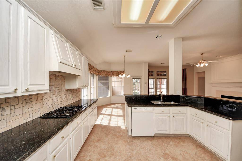 This bright and spacious kitchen features white cabinetry, granite countertops, and a modern tile backsplash. It includes a gas cooktop and a central island with a sink, opening to a sunlit breakfast nook and living area.