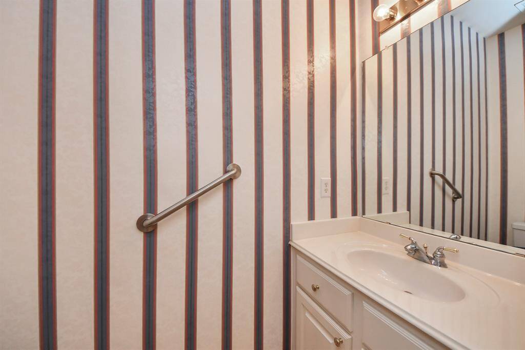 This bathroom features a single sink vanity with a mirror, striped wallpaper, and a wall-mounted handrail for support.