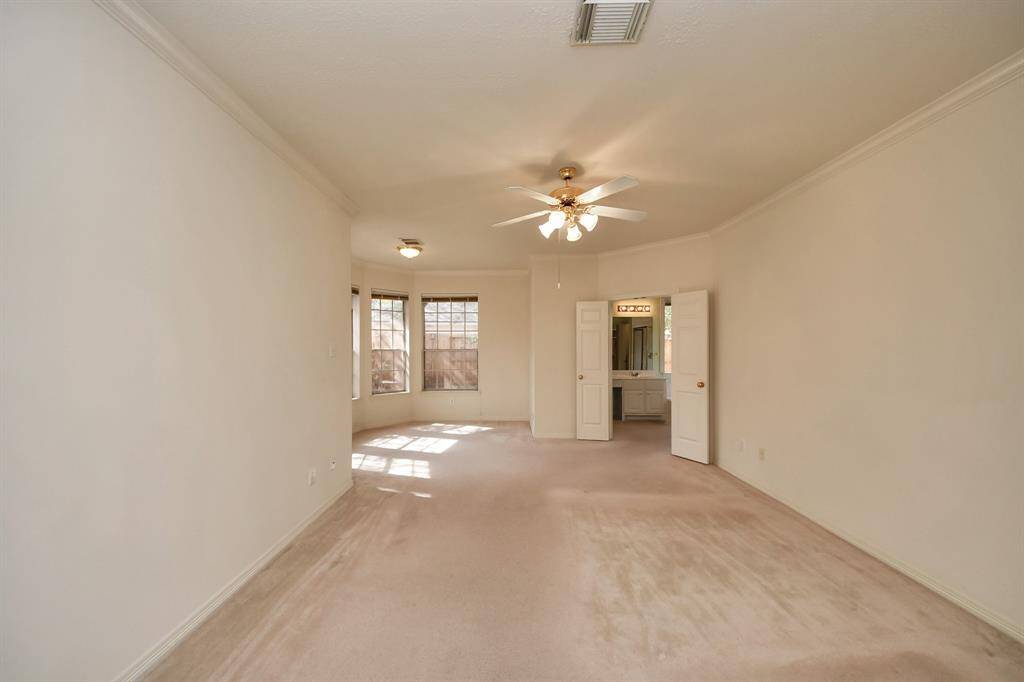 A spacious primary bedroom with neutral carpeting and walls, featuring a ceiling fan and ample natural light from large windows. It includes a small alcove and an open doorway leading to a bathroom with double doors, ideal for a cozy yet functional living space.