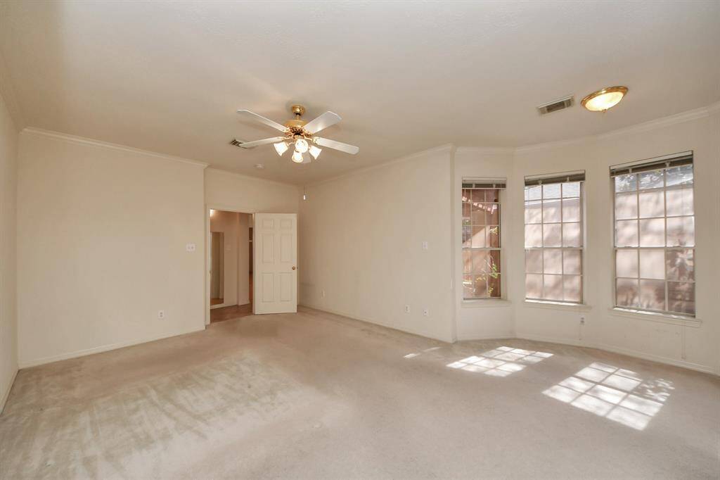 This spacious primary bedroom features neutral carpeting, a ceiling fan with lights, and ample natural light from large windows. The room has a doorway leading to another area, offering a cozy and inviting atmosphere, perfect for relaxation or entertaining.