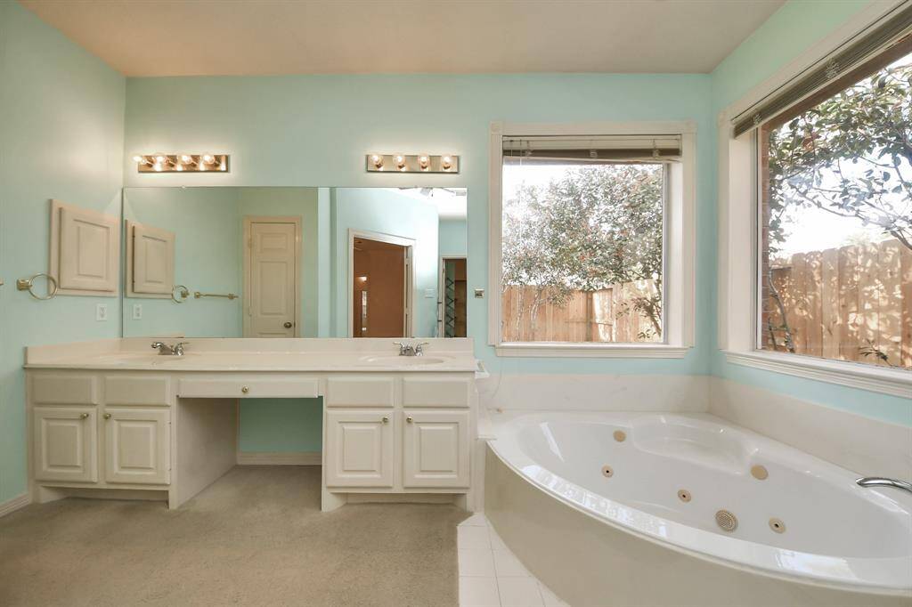 This primary bathroom features a spacious layout with a large double vanity, ample counter space, and under-sink storage. It includes a luxurious corner jacuzzi tub near large windows that allow natural light, creating a bright and inviting atmosphere.