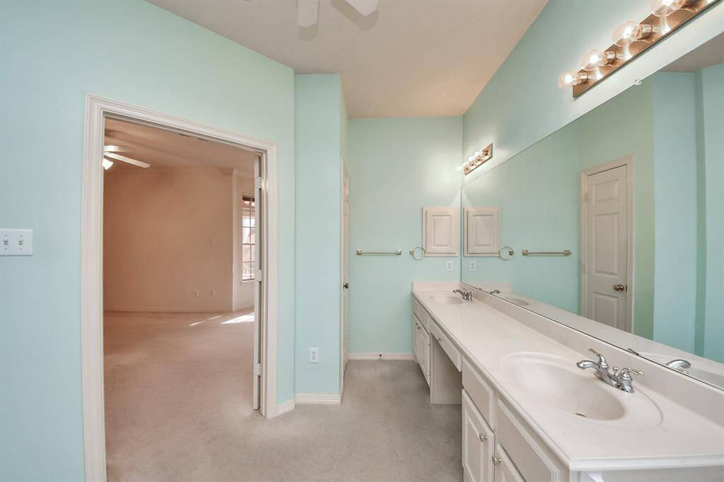 This photo shows a spacious bathroom with a double vanity featuring ample storage. The room is painted in a soft turquoise color and includes large mirrors and bright lighting. A doorway leads to an adjoining carpeted room with a ceiling fan and window.