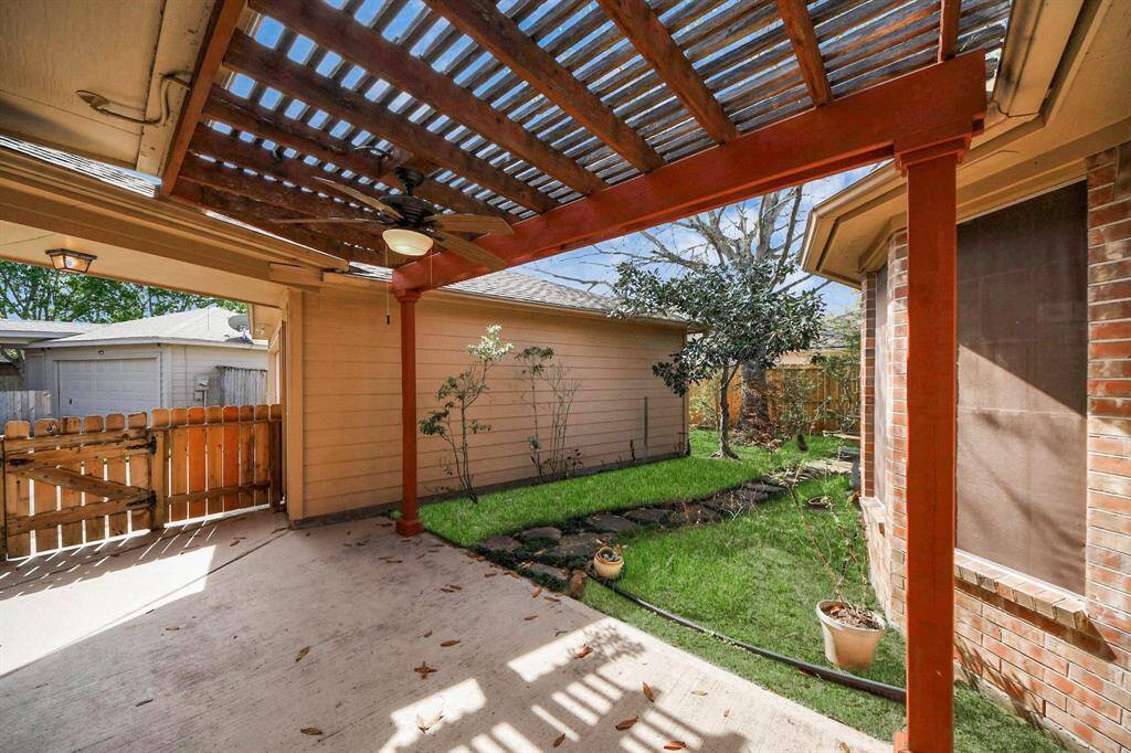 This photo showcases a cozy backyard patio with a wooden pergola, ceiling fan, and a small lawn area. It offers a private, shaded space ideal for relaxation or outdoor dining.