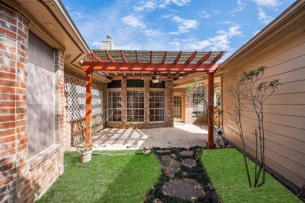 This photo showcases a cozy outdoor patio area with a wooden pergola, providing partial shade. The space is flanked by a mix of brick and siding walls, with a pathway leading through a small, well-maintained grassy area. Large windows offer a view into the home, enhancing the sense of openness and light. Ideal for relaxation or entertaining.