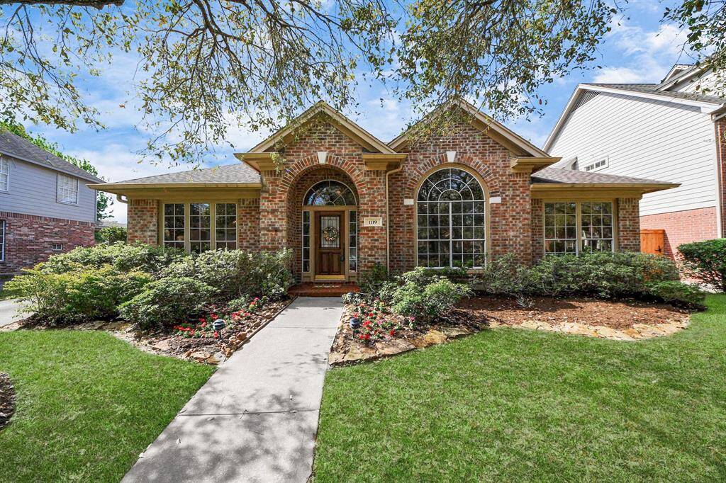 This charming brick home features a welcoming arched entryway, large windows, and well-maintained landscaping with lush greenery and colorful flowers, creating great curb appeal in a serene neighborhood setting.