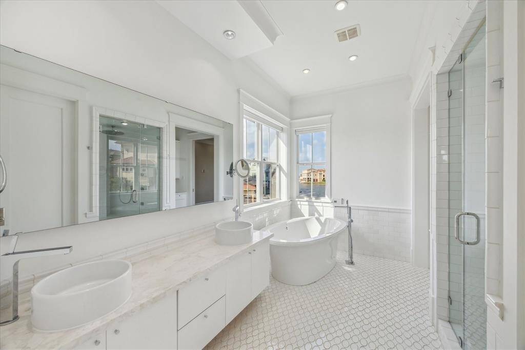 The primary bathroom is a bright, spa-inspired retreat featuring a luxurious soaking tub positioned beneath a bay window with serene water views. Crisp white finishes and sleek design elements enhance the airy, tranquil feel. Dual vessel sinks sit atop a marble vanity with abundant counter space, while a spacious glass-enclosed shower adds to the modern elegance. Recessed lighting and subtle architectural details complete this clean, calming oasis