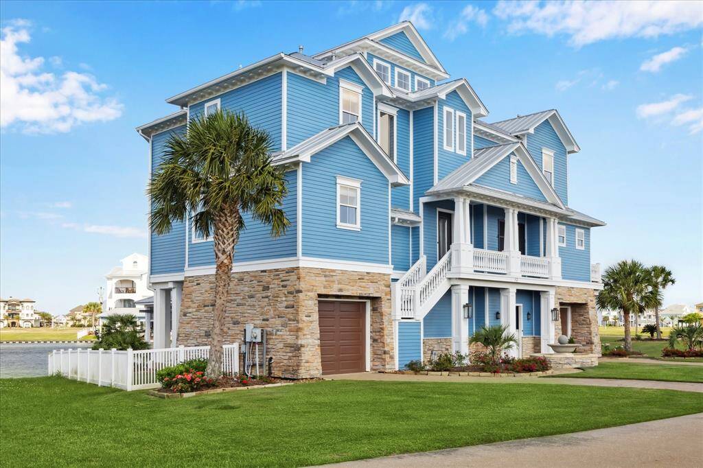 This stunning coastal home features classic blue Hardie siding with crisp white trim, accented by a natural stone for added texture and elegance. Its elevated design creates architectural interest and maximizing natural light. A broad front staircase leads to a covered entry porch, while palm trees and vibrant landscaping enhance the tropical, waterfront setting. The home’s position offers serene views of the lagoon, with easy access to the Harborwalk Marina Channel and West Galveston Bay