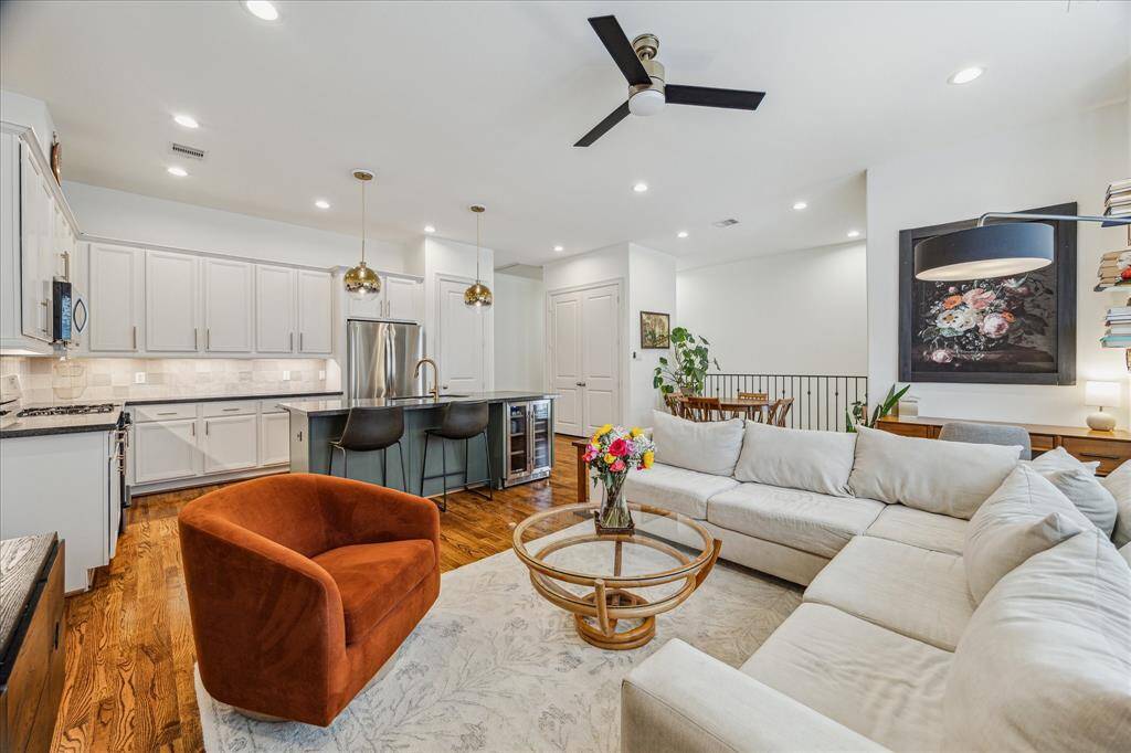 Bright and welcoming living room featuring warm hardwood flooring, recessed LED lighting, and neutral window coverings that complement any décor. The open layout seamlessly connects to the kitchen and breakfast room, creating an ideal space for both relaxing and entertaining.