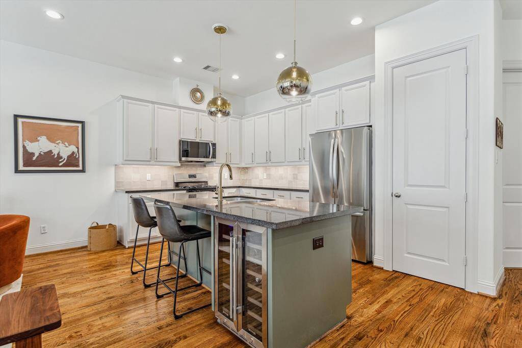 This stunning kitchen features designer-painted wood cabinetry accented by elegant champagne bronze hardware and a matching faucet. Under-cabinet lighting highlights the grey ceramic tile backsplash, while warm hardwood flooring adds a touch of richness and warmth to the space.