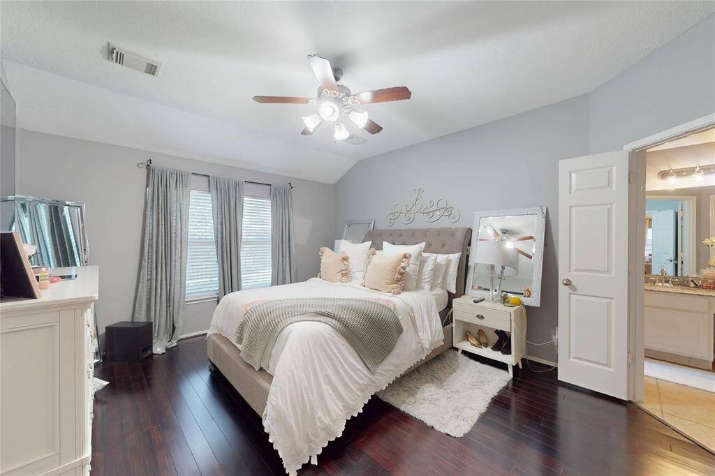 This primary bedroom offers a cozy and elegant atmosphere with dark hardwood floors and soft gray walls. It features a stylish ceiling fan, large windows with natural light, and an en-suite.