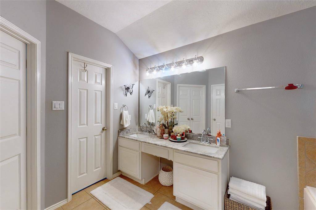 This primary bathroom en-suite features a double vanity with a large mirror and good lighting. It has a neutral color palette with gray walls and white cabinetry, providing a clean and modern look. There is additional storage space and decorative elements, adding to the room's functionality and appeal.