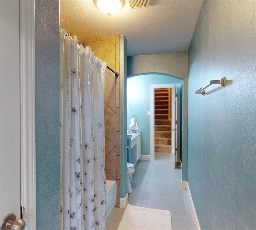 This bathroom features a cozy layout with a shower-tub combo, blue walls, and a white vanity. A hallway with stairs is visible in the background, adding a unique architectural touch.