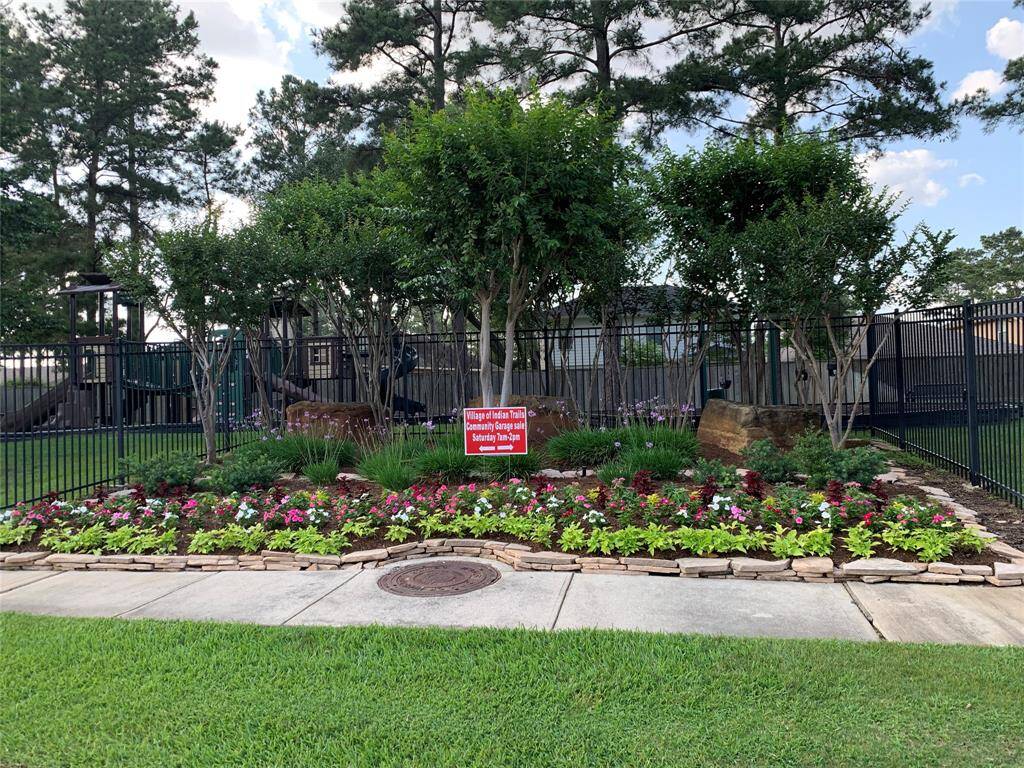Across the street, a landscaped garden area with colorful flowers and neatly arranged stone borders, set in front of a fenced playground. Tall trees provide shade, creating a welcoming community space.