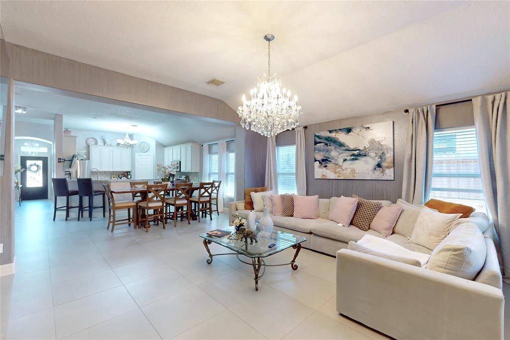 This spacious open-concept living area features a modern design, elegant chandelier, and neutral tile flooring. The adjoining kitchen and dining area offer ample natural light, with white cabinetry and spacious dining area creating a welcoming and functional space for entertaining.