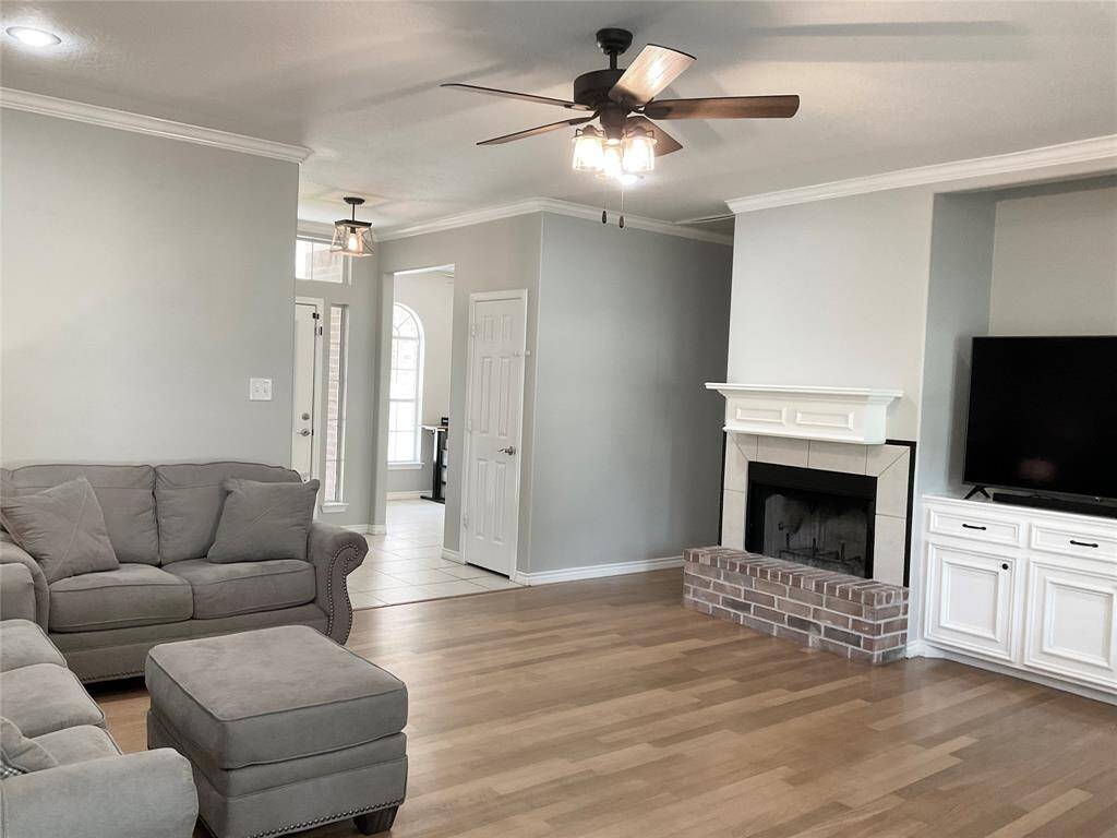 This cozy living room features light wood flooring, a brick, wood burning fireplace with a white mantel, built-in cabinetry, a ceiling fan, and soft, neutral wall colors. The entryway is visible with ample natural light from large windows.  Notice the home office upfront with 3 beautiful windows.