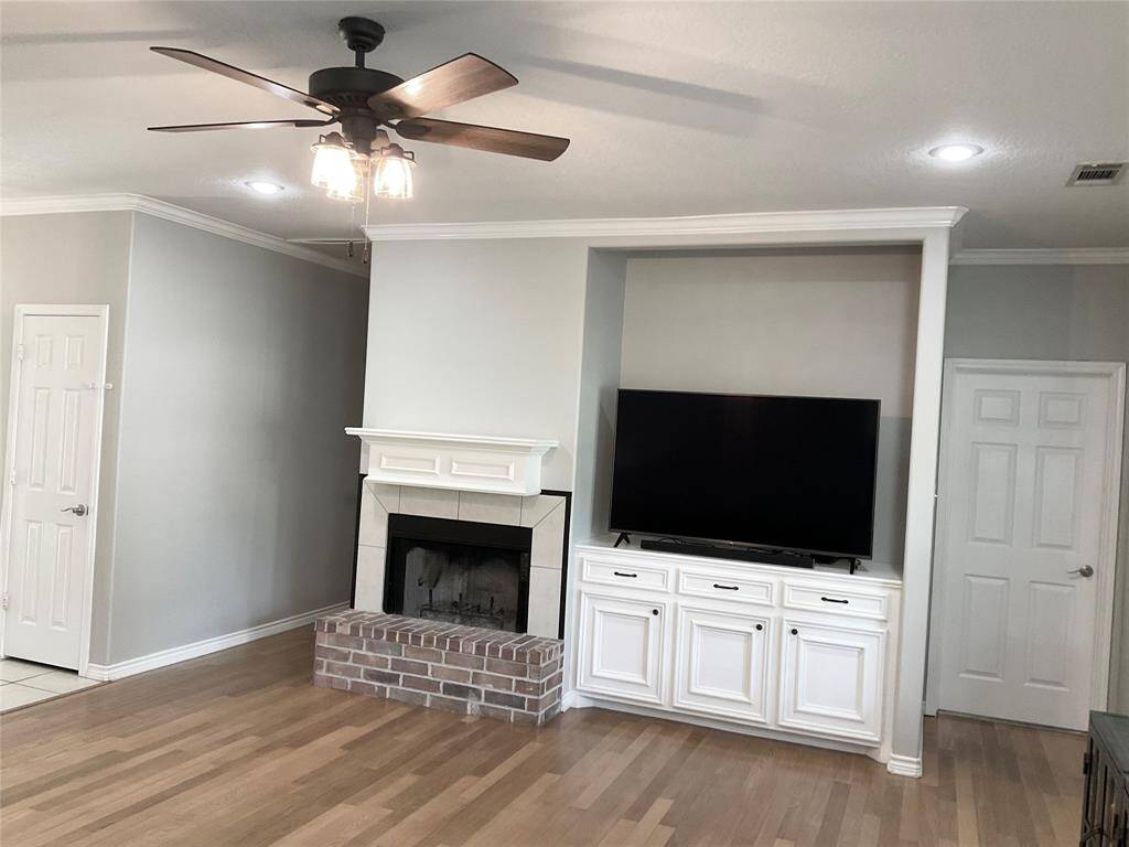 This living room features a cozy brick fireplace, built-in cabinetry with a TV, hardwood floors, and a ceiling fan with overhead lighting, creating a warm and inviting space.  Lush views of the back yard are to the right.