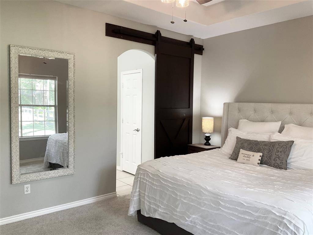 The primary bedroom features a cozy atmosphere with a soft color palette. It includes a large window for natural light, a stylish barn door, and a wall mirror. The room is spacious and accommodates a king size bed with a view of the beautiful back yard.