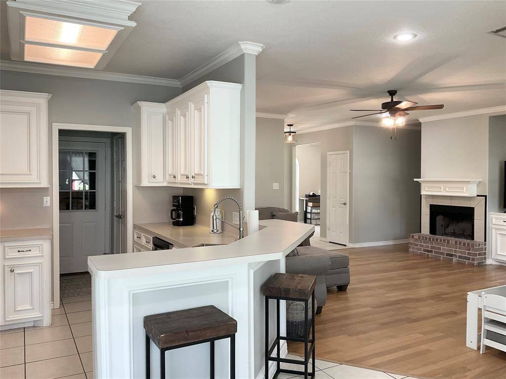Showcasing a bright, open-concept living area with a modern kitchen featuring white cabinetry and a breakfast bar. The space flows into a cozy living room with a ceiling fan, hardwood floors, and a brick fireplace, creating a warm and inviting atmosphere.