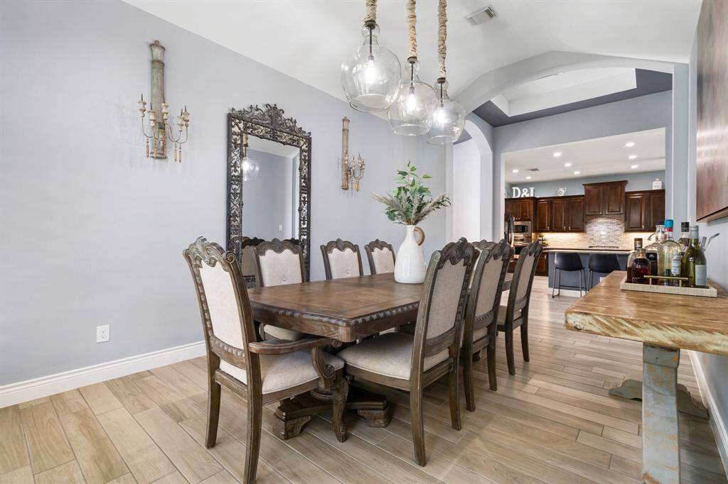 The dining room features elegant wooden flooring and a large, ornate dining table with upholstered chairs. A decorative mirror and wall sconces add sophistication, while a modern light fixture illuminates the space. The room opens to a spacious kitchen with dark wood cabinetry and a light backsplash.