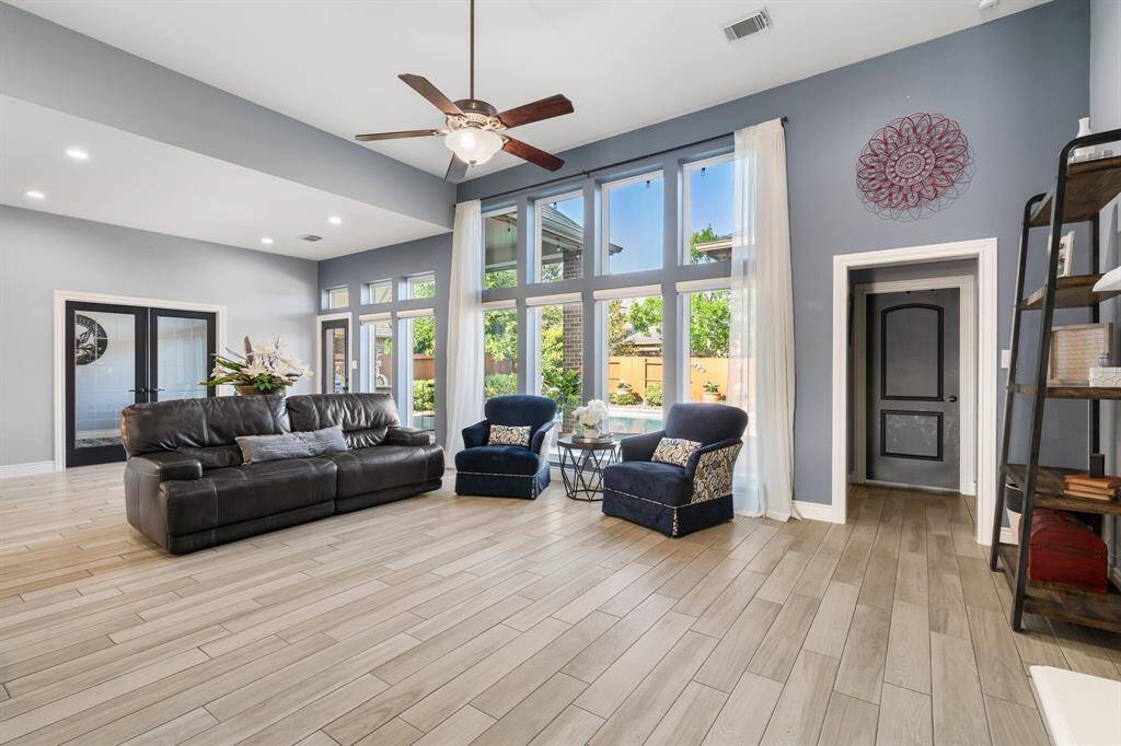 This spacious living room features high ceilings and abundant natural light from large windows. It includes a comfortable seating area with a leather sofa and two armchairs, modern wood-look tile flooring, and stylish decor. Perfect for family gatherings or relaxation.