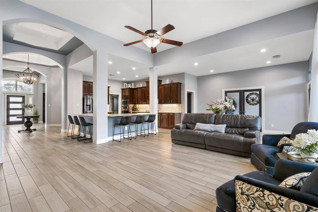 This spacious open-concept living area features elegant wood-look tile flooring, a modern kitchen with bar seating, and a cozy seating area with a leather sofa. The room is well-lit with recessed lighting, and the entrance showcases a stylish chandelier and contemporary double doors.