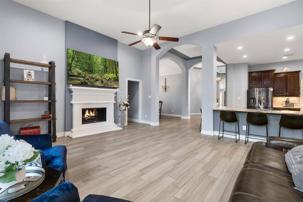 A spacious living area with a cozy fireplace, modern ceiling fan, and open layout. It features wood-look tile flooring, a mounted TV, and flows into a sleek kitchen with dark cabinetry and a breakfast bar, perfect for entertaining.