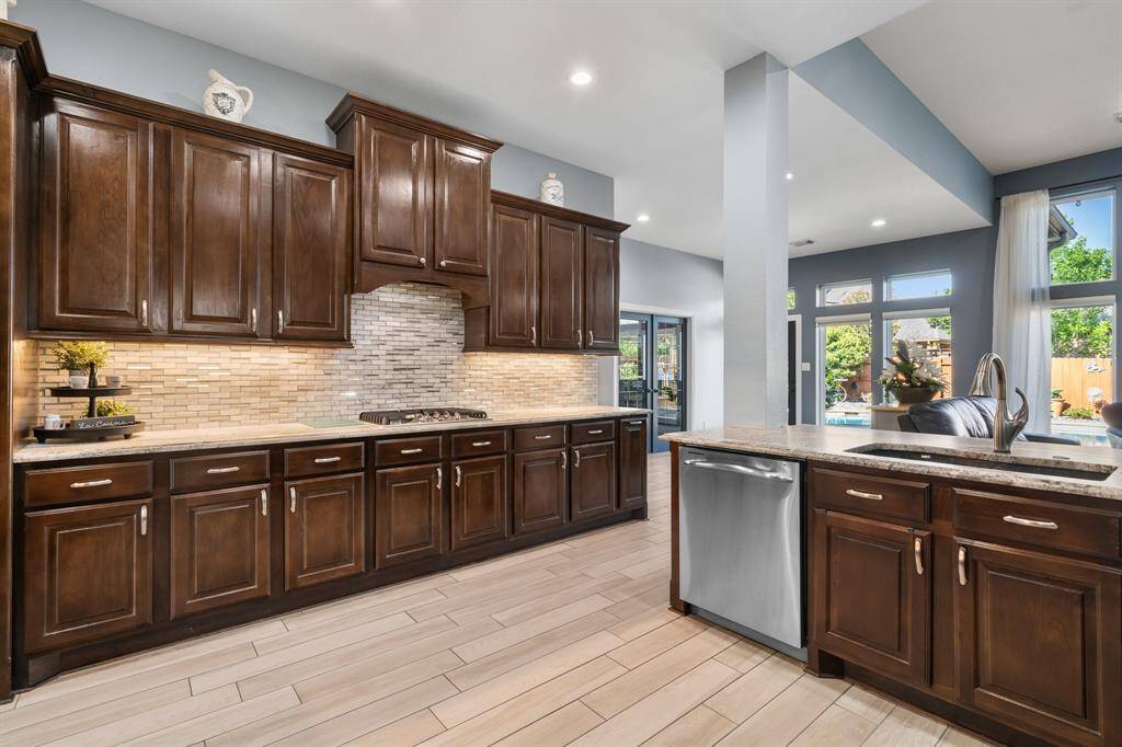 This kitchen features elegant dark wood cabinets, a modern tile backsplash, and stainless steel appliances. It opens to a bright living area with large windows offering views of the patio and pool.