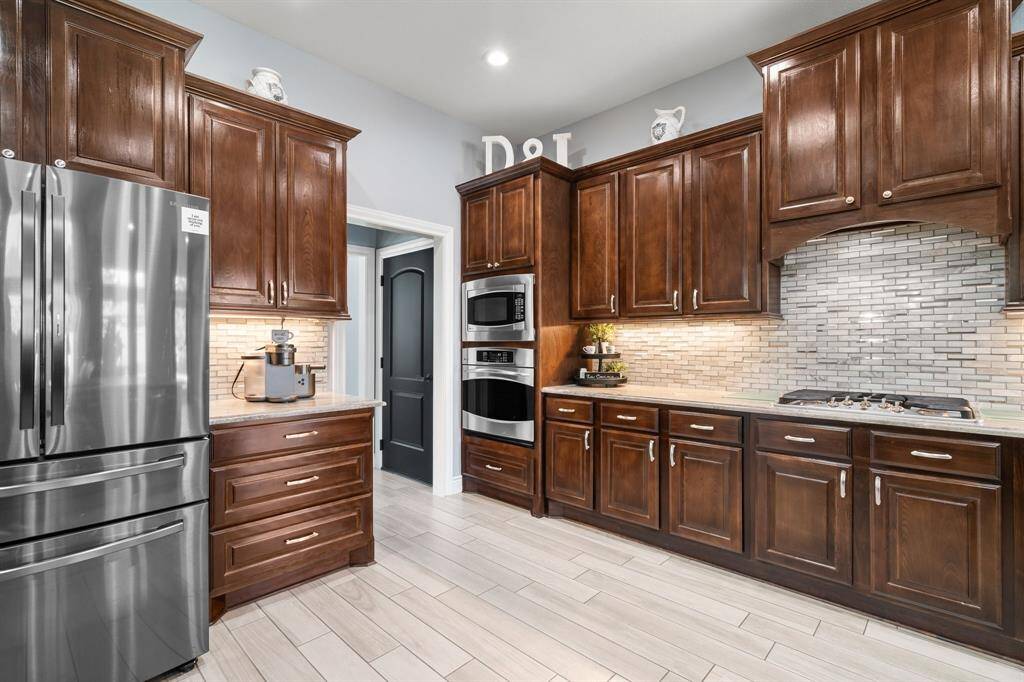 This kitchen features elegant dark wood cabinets, stainless steel appliances, a modern tile backsplash, and light-colored flooring. It offers ample storage and countertop space, with a sleek and contemporary design.