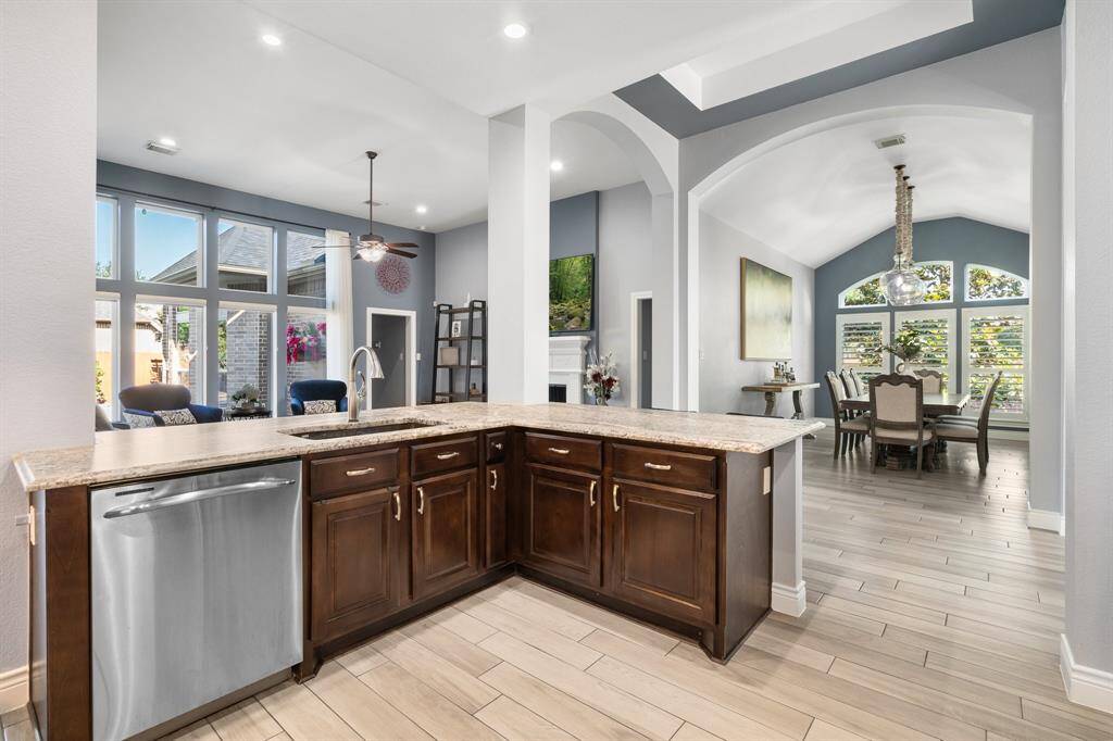 Explore this elegant kitchen and dining area with dark wood cabinets, a stone island, and stainless steel appliances. Large windows and a high ceiling with a chandelier fill the space with natural light, creating a warm and inviting atmosphere.