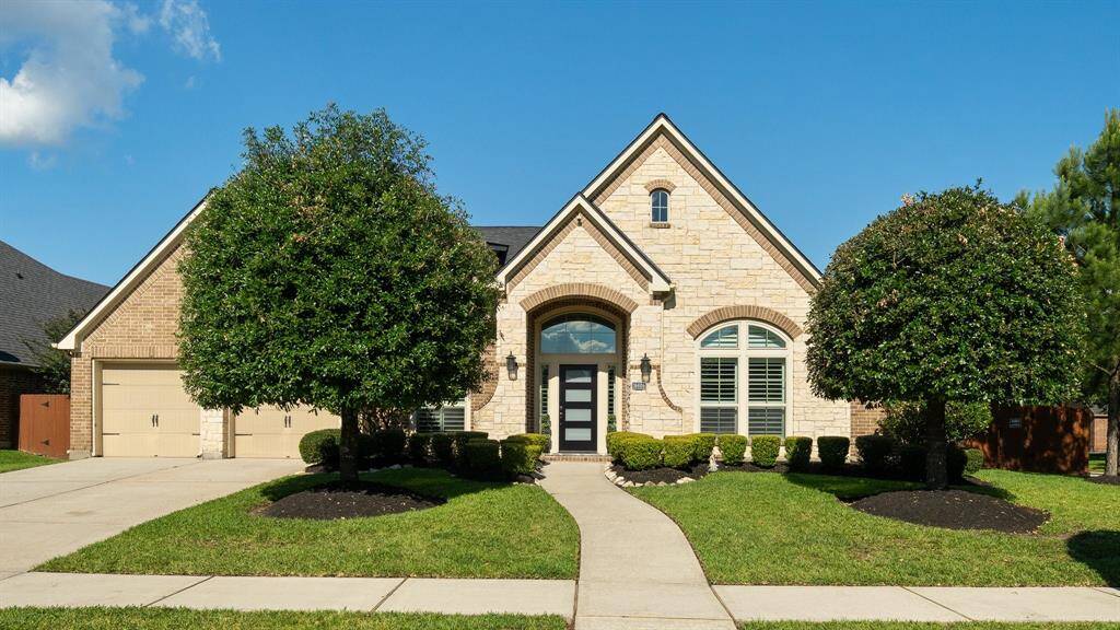 This charming home features a welcoming stone and brick exterior with a gabled roof, arched entryway, and well-manicured landscaping. The driveway leads to a two-car garage, and large windows provide plenty of natural light.