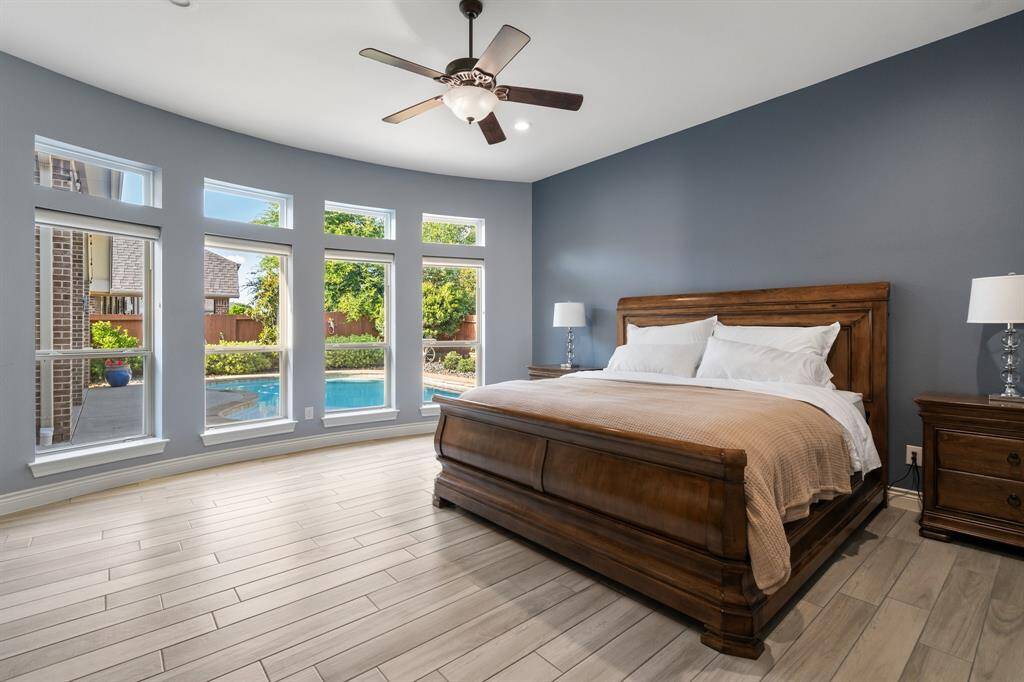 Experience luxury in this spacious primary bedroom featuring a large wooden bed, elegant gray walls, and wood-look tile flooring. Abundant natural light streams through multiple windows, offering stunning views of the private pool and landscaped backyard. A stylish ceiling fan enhances comfort.