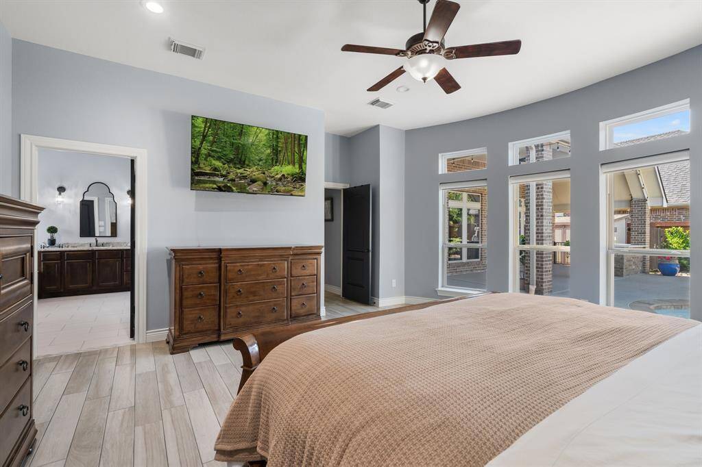 Experience the charm of this primary bedroom with expansive windows, a ceiling fan, and a mounted TV. The ensuite bathroom features dual sinks and elegant dark wood cabinetry. Neutral tones and wood-look flooring create a cozy, modern ambiance, while overlooking the patio and pool.
