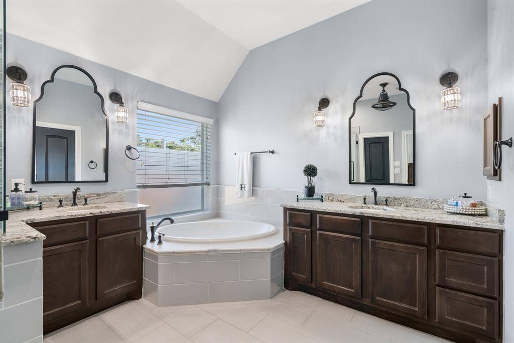 This elegant bathroom features dual vanities with granite countertops, stylish mirrors, and modern light fixtures. A large soaking tub is centrally located, complemented by natural light from a window with privacy blinds. The space offers a calm, contemporary design.
