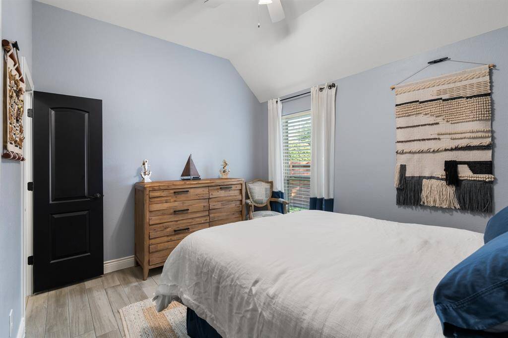 Cozy bedroom with gray walls, a vaulted ceiling, and natural light from a window. Features a wooden dresser, a stylish woven wall hanging, and a modern black door.