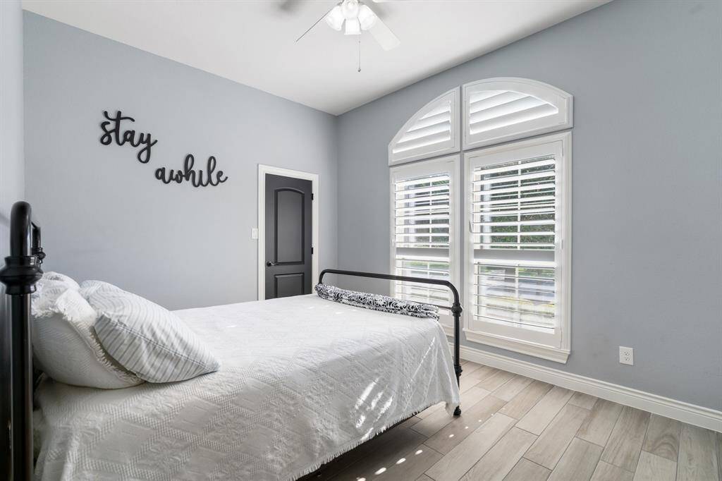 Bright bedroom with neutral tones features a bed, decorative wall art, and large arched windows with shutters, allowing natural light to fill the space.