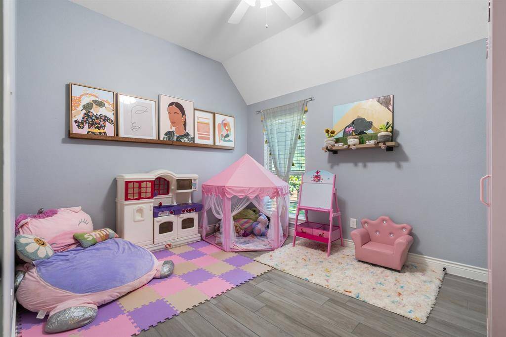 This spacious third bedroom has been transformed into a cozy children's playroom, featuring soft gray walls and wood-look tile flooring. It includes a play kitchen, pink play tent, and a small pink chair. Bright artwork and toys add charm, while a window fills the room with natural light.