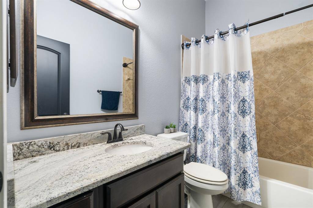 This bathroom features a stylish countertop with a dark wood vanity, a large framed mirror, and a shower-tub combo with a patterned curtain. The neutral color scheme and modern fixtures create a welcoming and functional space.
