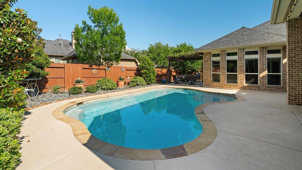 Discover a serene backyard oasis featuring a pristine swimming pool, expansive concrete deck, and lush greenery. Enjoy privacy with a wooden fence and relax in the cozy seating area beneath a pergola attached to the charming brick house—ideal for entertaining and unwinding.