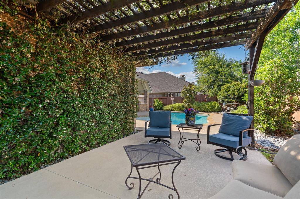 Showcases a charming patio with cozy seating beneath a pergola draped in lush greenery. Enjoy views of a sparkling pool and beautifully landscaped garden, creating your perfect backyard oasis.