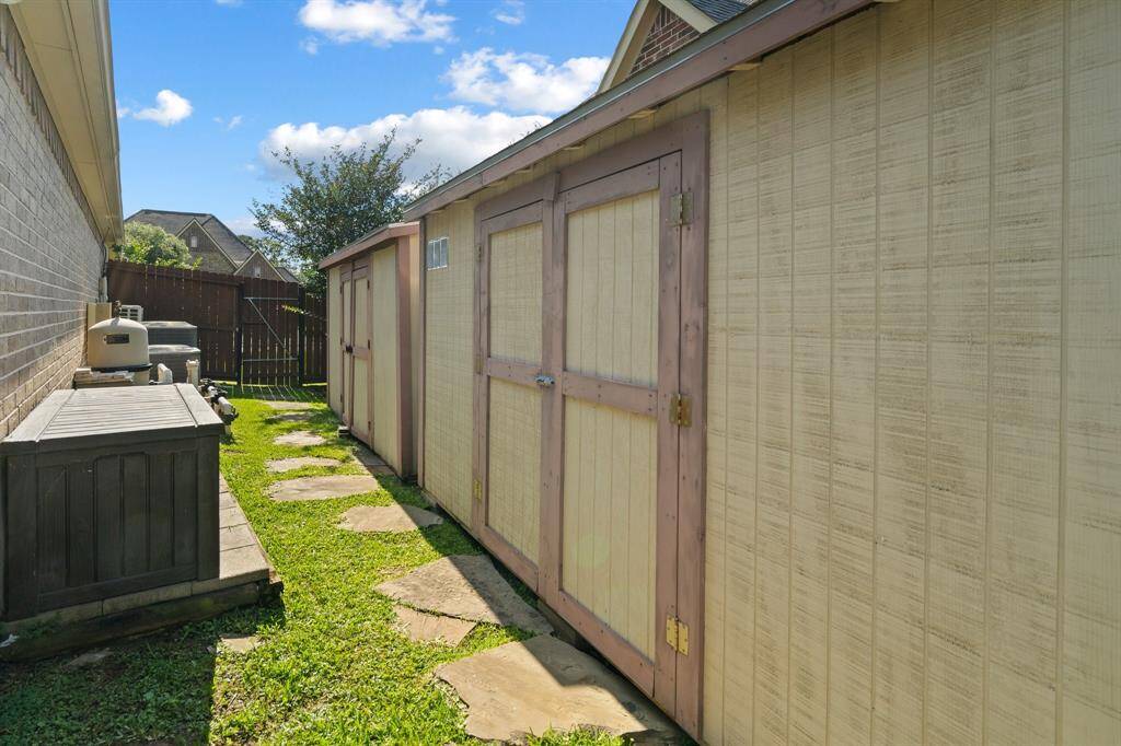 Charming side yard featuring two wooden shed and stone pathway, ideal for storage or a workspace, with a lush grassy area and a brick wall backdrop.