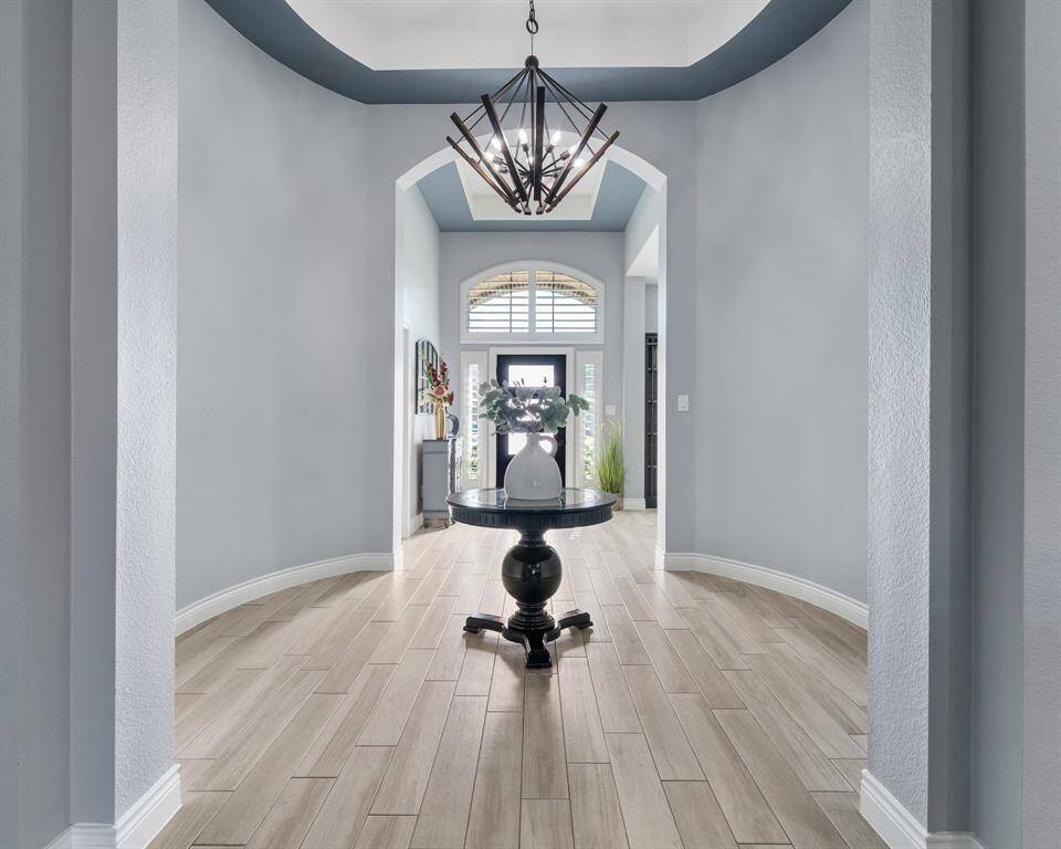 This photo showcases an elegant entryway with light gray walls, wood-look tile flooring, and a decorative round table with a vase centerpiece. A stylish chandelier hangs from the ceiling, and the space is well-lit with natural light from the front door's window panels.
