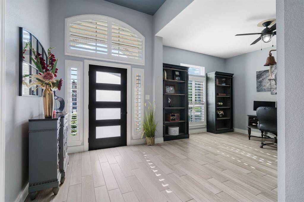 Bright entryway with modern black door, large windows, and stylish tile flooring. Adjacent room features shelving, a ceiling fan, and ample natural light, ideal for a home office.