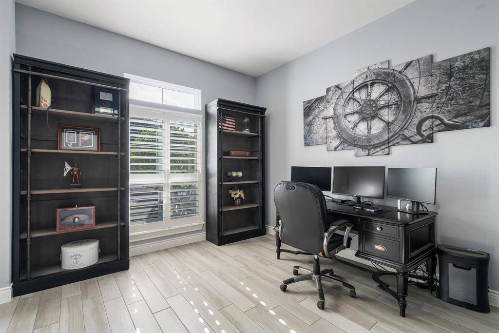 This photo shows a bright and inviting home office with wood-look tile flooring, bookshelves, and a large window with plantation shutters. It features a stylish desk setup with multiple monitors and nautical-themed wall art, creating a functional and elegant workspace.