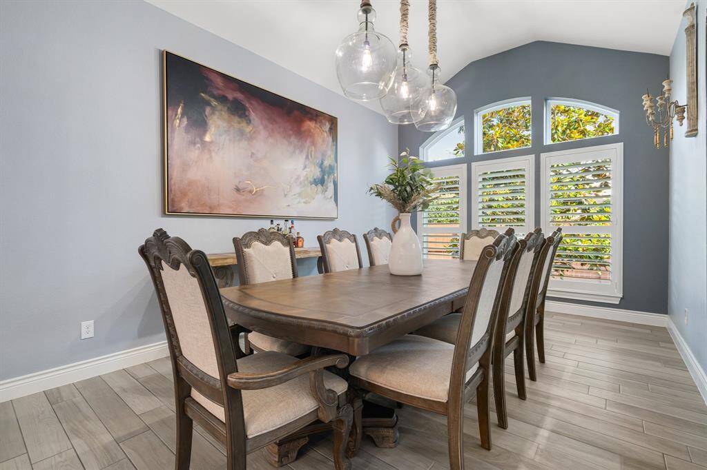 This dining room features elegant wooden furniture with a large table and upholstered chairs. It has a high ceiling with a modern chandelier, a large abstract painting, and a series of arched windows with plantation shutters, providing ample natural light.