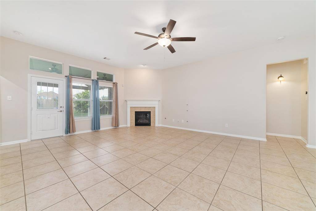 Spacious living room with tiled floors, a ceiling fan, and a cozy fireplace. Large windows provide natural light, and there's an adjacent entry door. Neutral walls offer a clean, modern feel.
