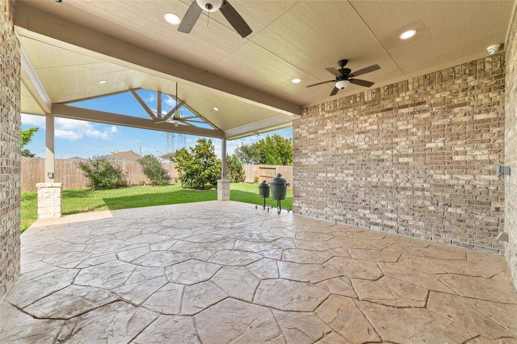 This photo features a spacious covered patio with a stone-textured floor, ceiling fans, and a brick wall. It opens to a green backyard, perfect for outdoor gatherings and relaxation.
