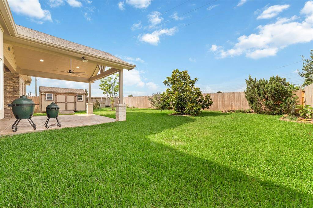 Spacious backyard with well-maintained lawn, covered patio with ceiling fan, and two grills. There's a wooden storage shed and privacy fence, perfect for outdoor relaxation and entertaining.