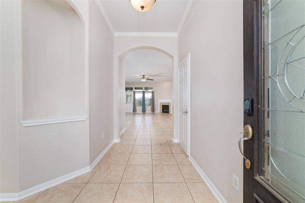 This photo shows a bright entryway with tile flooring, leading to a living area. It features high ceilings with an archway, neutral walls, and a glimpse of a fireplace and large windows in the background.