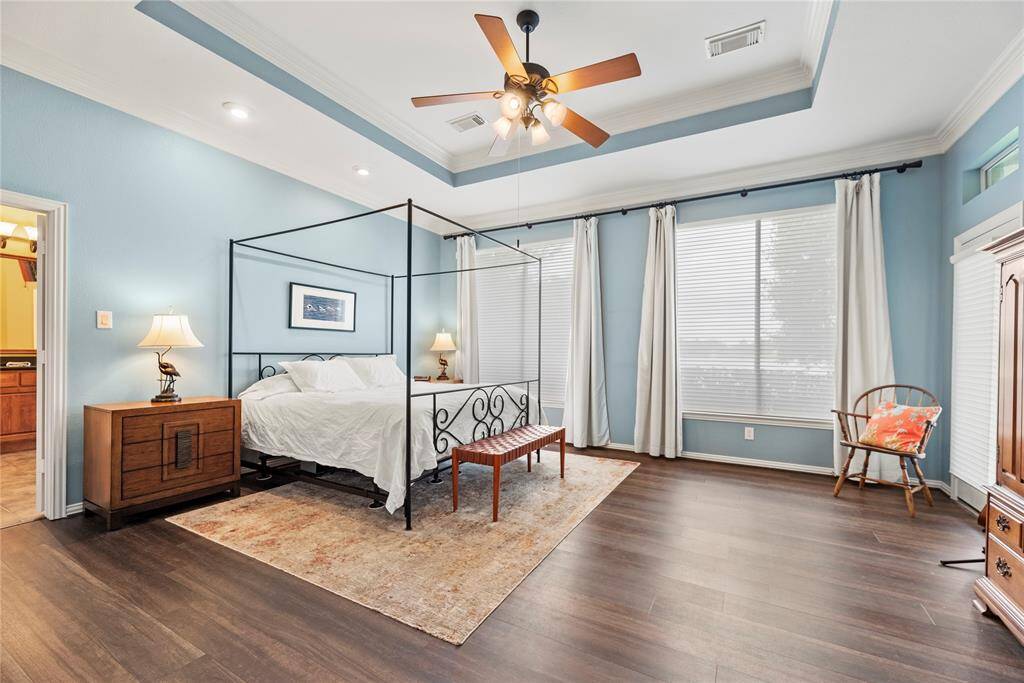Spacious primary bedroom with a rich wood flooring, and a tray ceiling with a ceiling fan, and ample natural light from large windows.